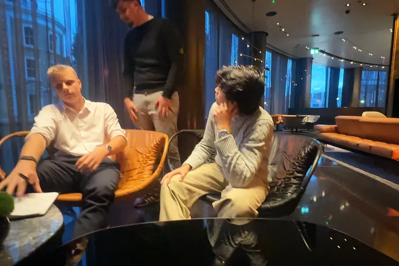 Three young founders talking by a tall window, two in armchairs and one standing